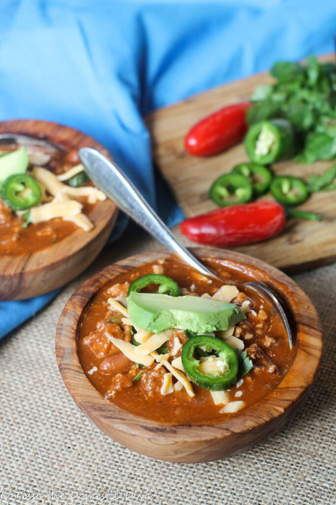 two hand carved wooden bowls filled with a tomato , bean, and ground meat stew with fresh avocado and jalapeño slices