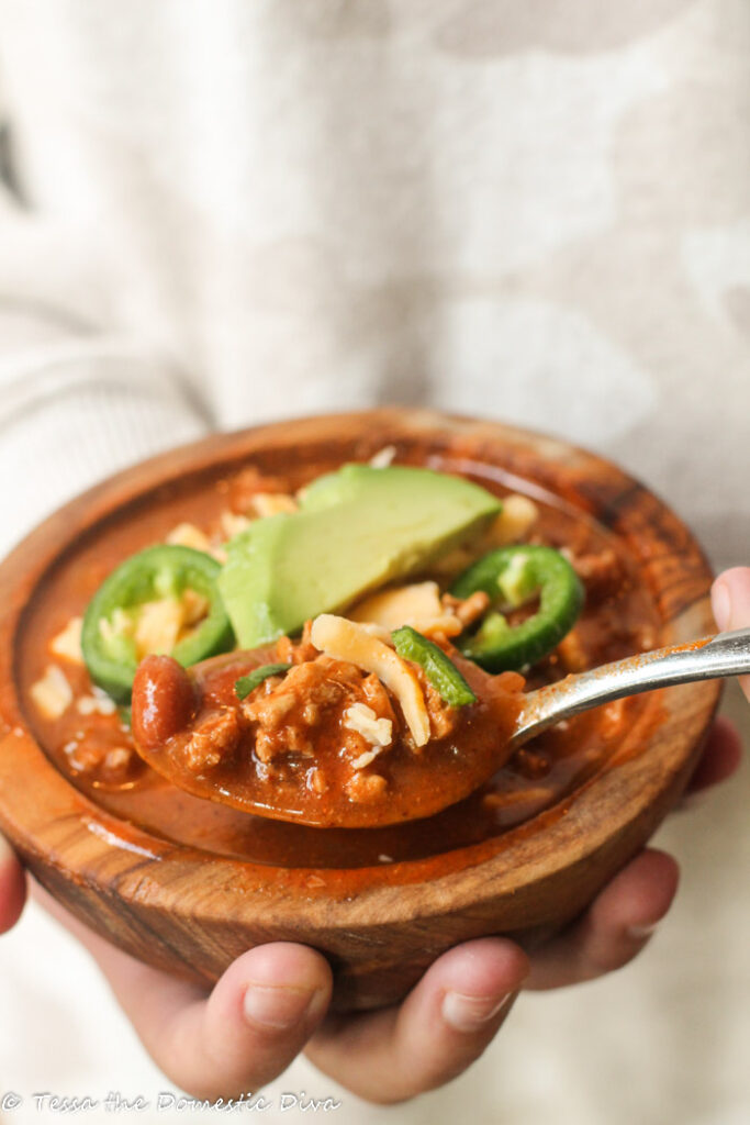 hand held wooden bowl filled with chili and topped with frash avocado, cilantro, and cheese