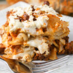 a square of noodle casserole layered with a creamy white cheese sauce and red meat sauce atop a white plate