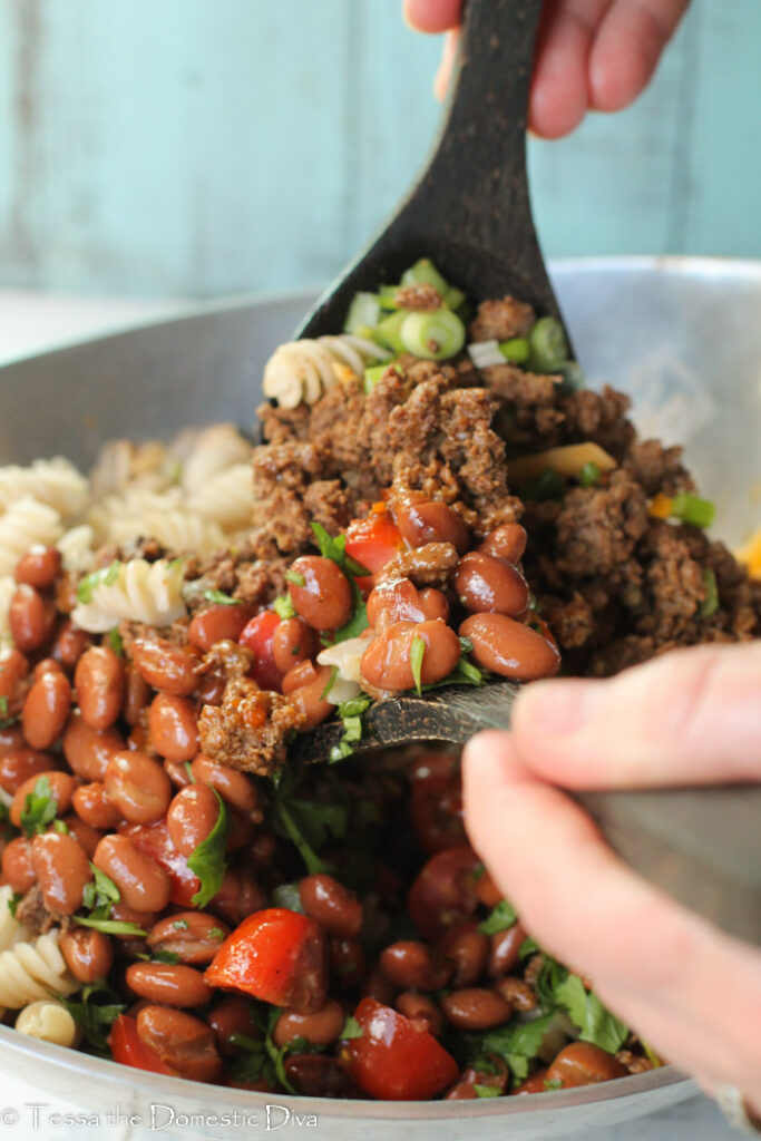 two hand help wooden spoons tossing the ingredients for a tex-mex pasta salad in a silver bowl