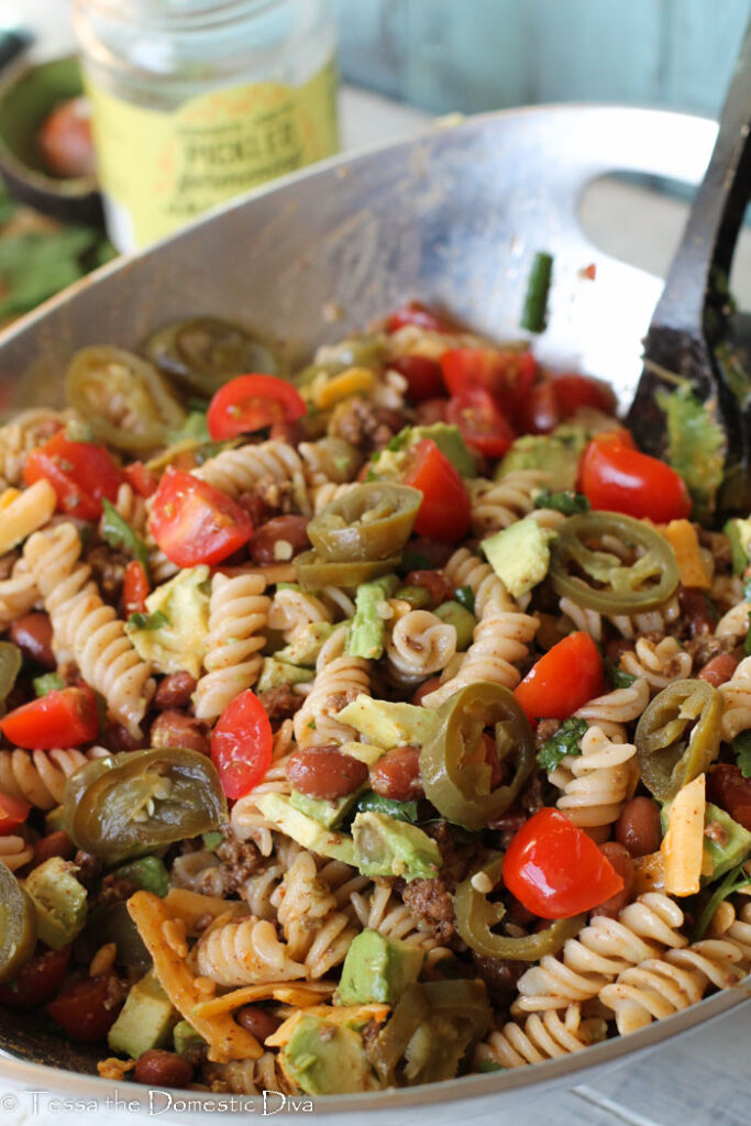 a silver bowl filled with fusilli noodles with avocado, tomatoes, cilantro, taco meat, shredded cheese, and pinto beans