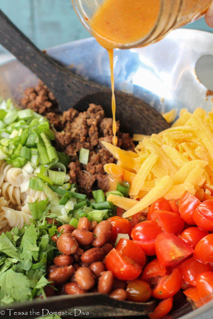 a chipotle vinaigrette being poured over the top of pinto beans, fusilli noodles, cheese, tomatoes, taco meat, cilantro, and green onions