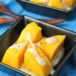 two square black dishes with peeled and cut chunks of thai mangoes with a drizzles of coconut sauce and sesame seeds