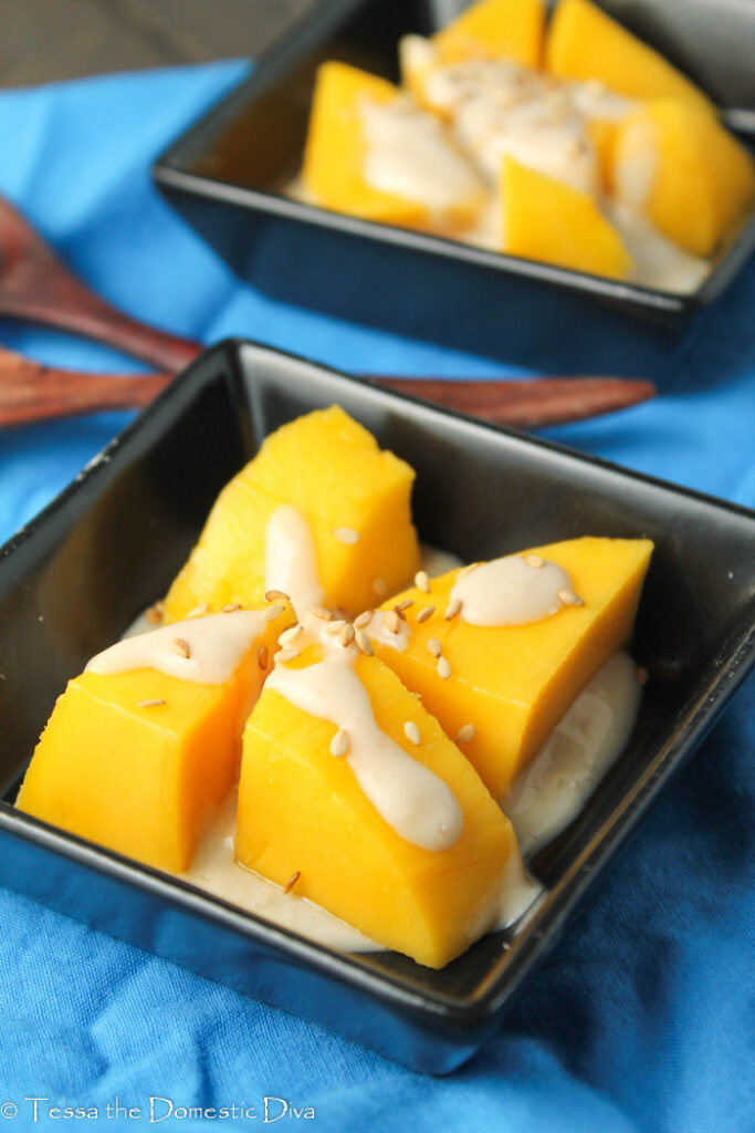 two square black dishes with peeled and cut chunks of Thai mangoes with a drizzles of coconut sauce and sesame seeds