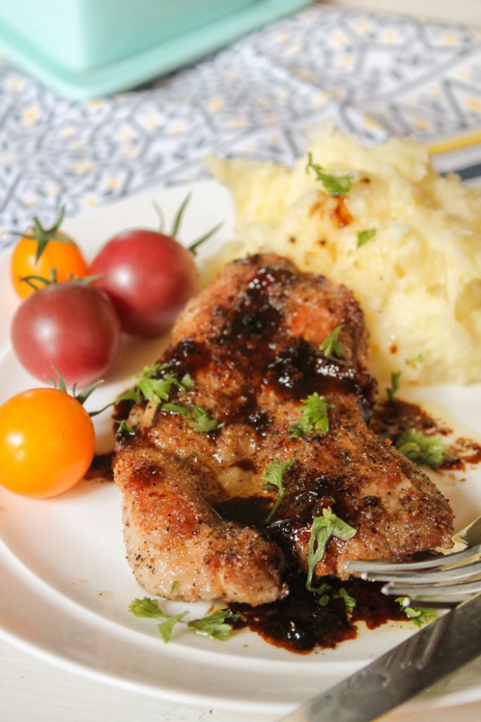 pork medallion seared in a skillet with a savory drizzle on a white plate with a mashed cauliflower and an heirloom cherry tomato side.