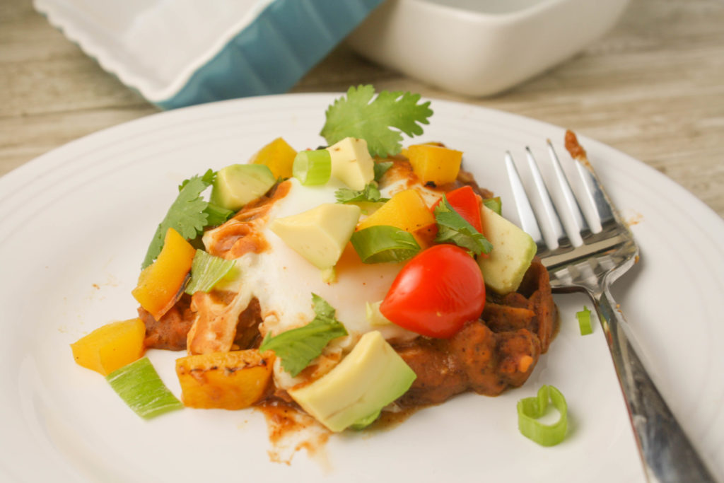 a white plate with a fork filled with refried bean and a molten poached egg with cilantro, tomato, and avocado garnishes