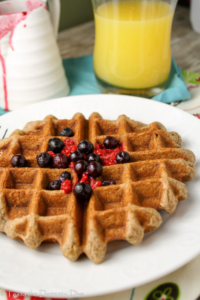 front view of a golden brown waffle with fresh berries, orange juice, and a pitcher of syrup