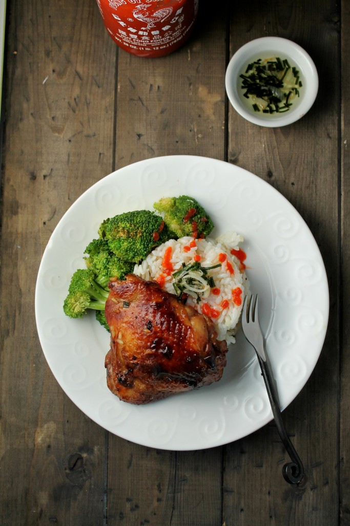 white plate of a golden gone in chicken thigh atop a mound of white rice with scallion oil, sriracha, steamed broccoli from overhead on a dark wooden table