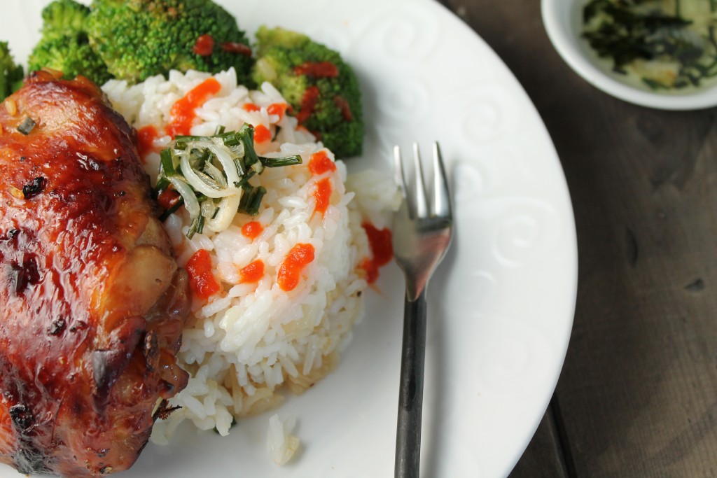close up of an asian roasted chicken thigh atop a serving of white rice with a drizzle of scallion oil and steamed broccoli