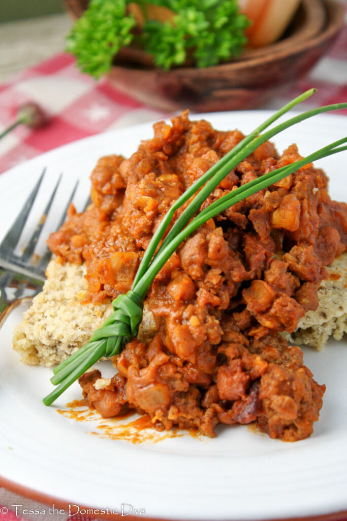 a white plate with corn bread topped with pinto beans, bacon, and ground beef in a barbecue sauce