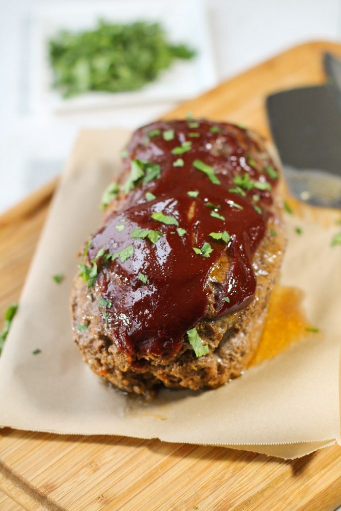 overhead shot of a whole meatloaf atop a bamboo cutting board slathered in a shiny tomato sauce with chopped parsley.