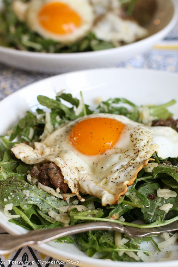 a white bowl with organic mixed grens and shaved sharpd cheese slices and topped with a fried egg and cooked breakfast sausage