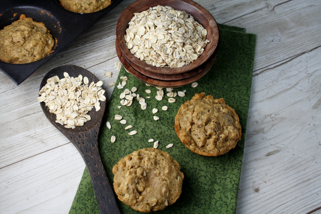 birdseye view of two oat studded muffins with a bowl of rolled gluten free oat in a wooden bowl atop a dark green cloth