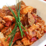 a square white bowl filled with a cajun rice, chicken, and sausage dish cooked in a tomato sauce