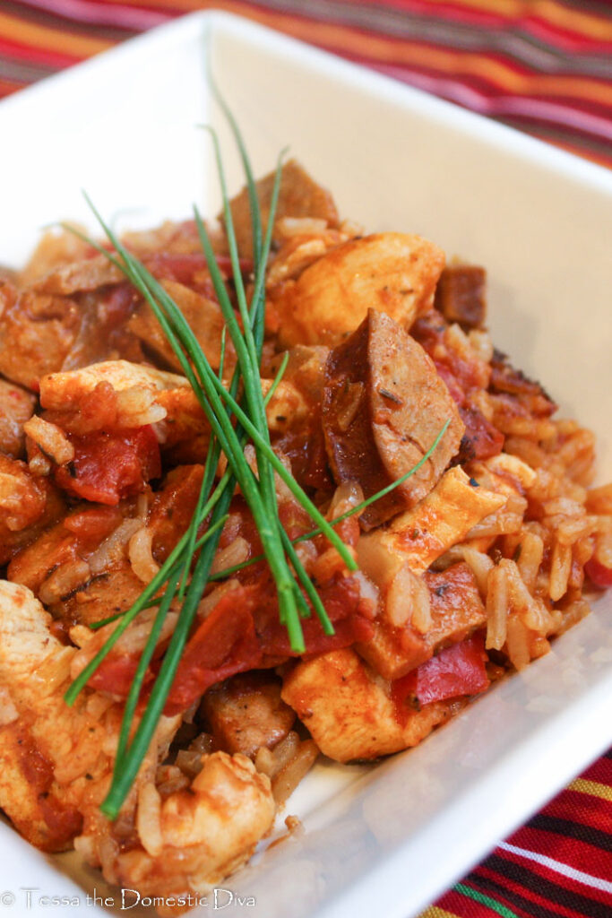 a square white bowl filled with a cajun rice, chicken, and sausage dish cooked in a tomato sauce