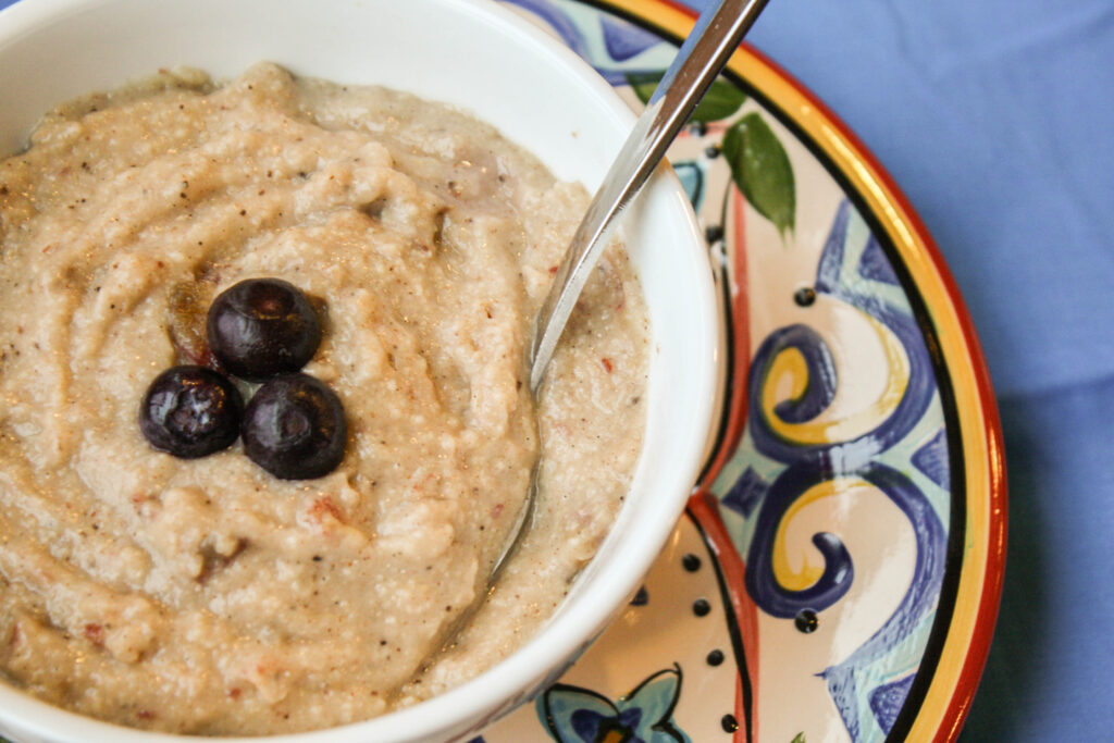 a white bowl filled with a creamy millet hot cereal topped with three blueberries atop a festive patterned plate