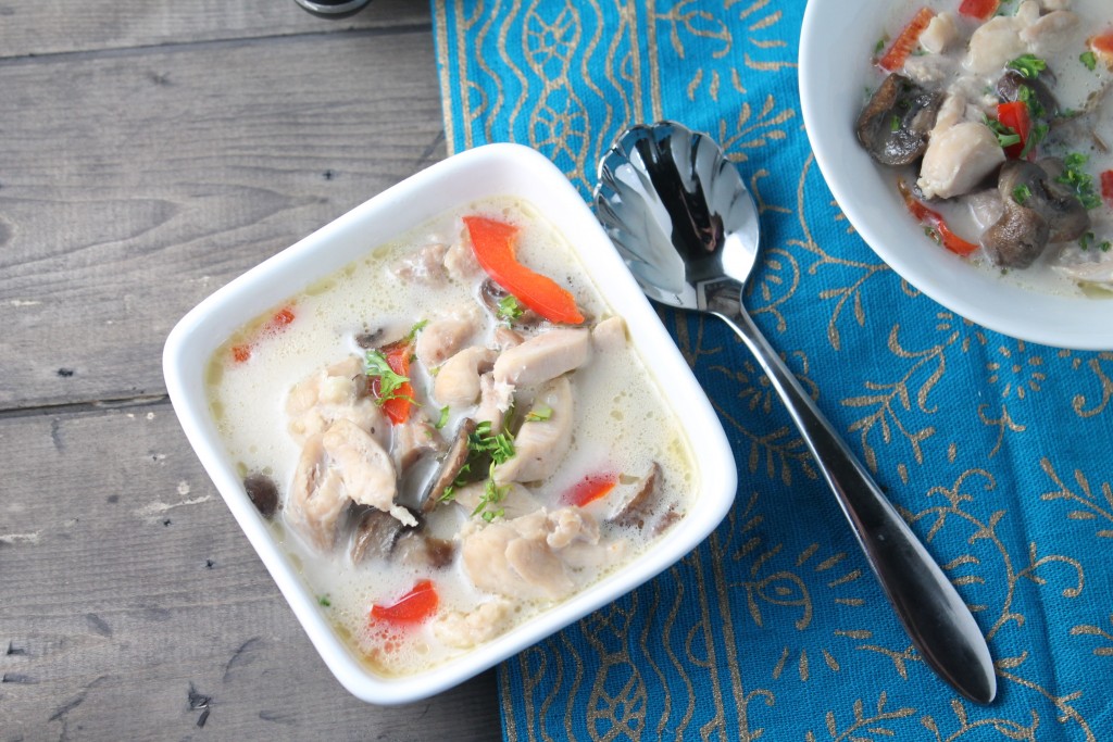 horizontal image of an overhead view of a square bowl of a coconut milk broth soup studded with tender bite sized pieces of chicken, fresh red bell pepper, mushrooms, and garnished with cilantro on a dark wooden surface