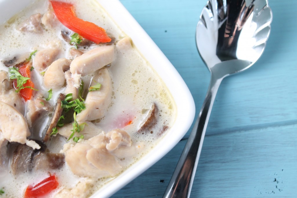 horizontal image of a white square bowl filled with a creamy coconu milk broth with boneless chicken thigh bite size pieces, red bell peppers, mushrooms, and a cilantro garnish on a distressed aqua wooden surface