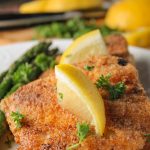 close up view of two pan fried breaded fish pieces on a white plat with lemon and parsley