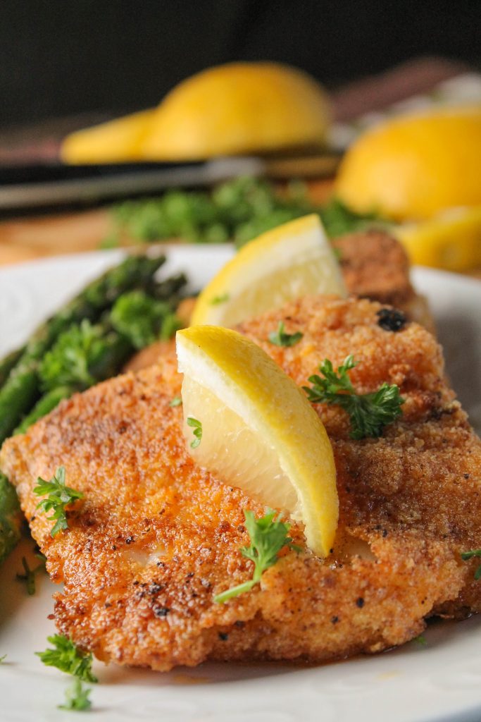 close up view of two pan fried breaded fish pieces on a white plat with lemon and parsley