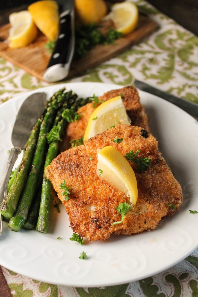 a white plate filed with two crispy breaded fish pieces and a side of asparagus with lemon and parsley