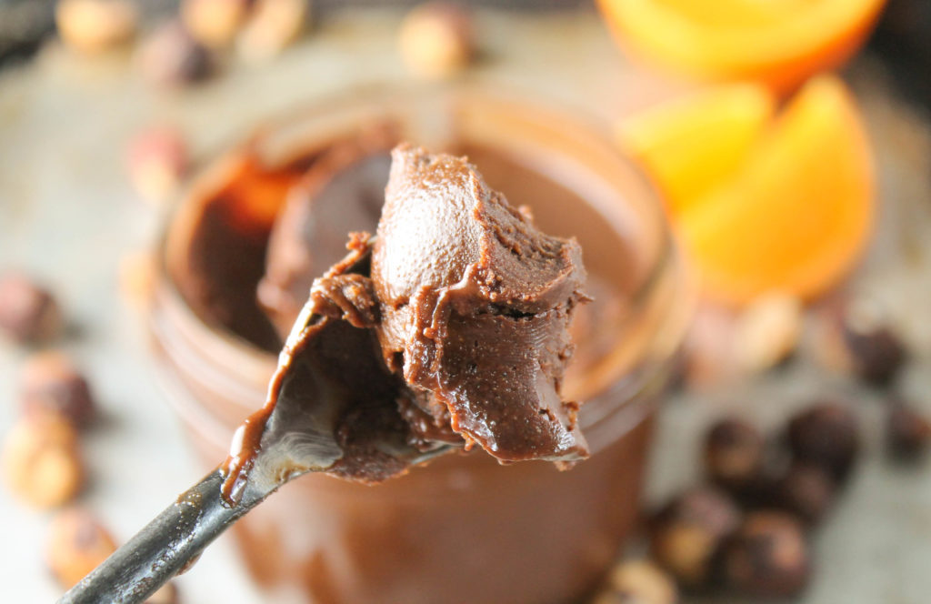 spoonful of nutella closeup with mason jar, orange slices, and roasted hazelnuts in foreground