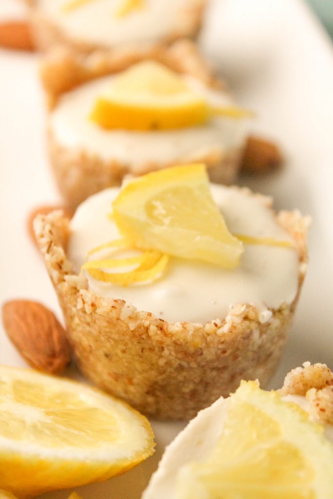 a row of lemon cheesecake tartlets in a raw nut crust with a garnish of fresh lemon slices on a white plate
