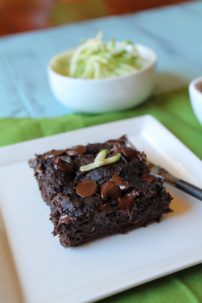 dark chocolate square of zucchini cake with melty chocolate chips and a a garnish of fresh zucchini on a white plate