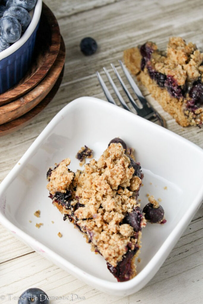 an oatmeal blueberry crumb bar triangle from overhead in a white square bowl with fresh blueberries scattered around on a light wood surface