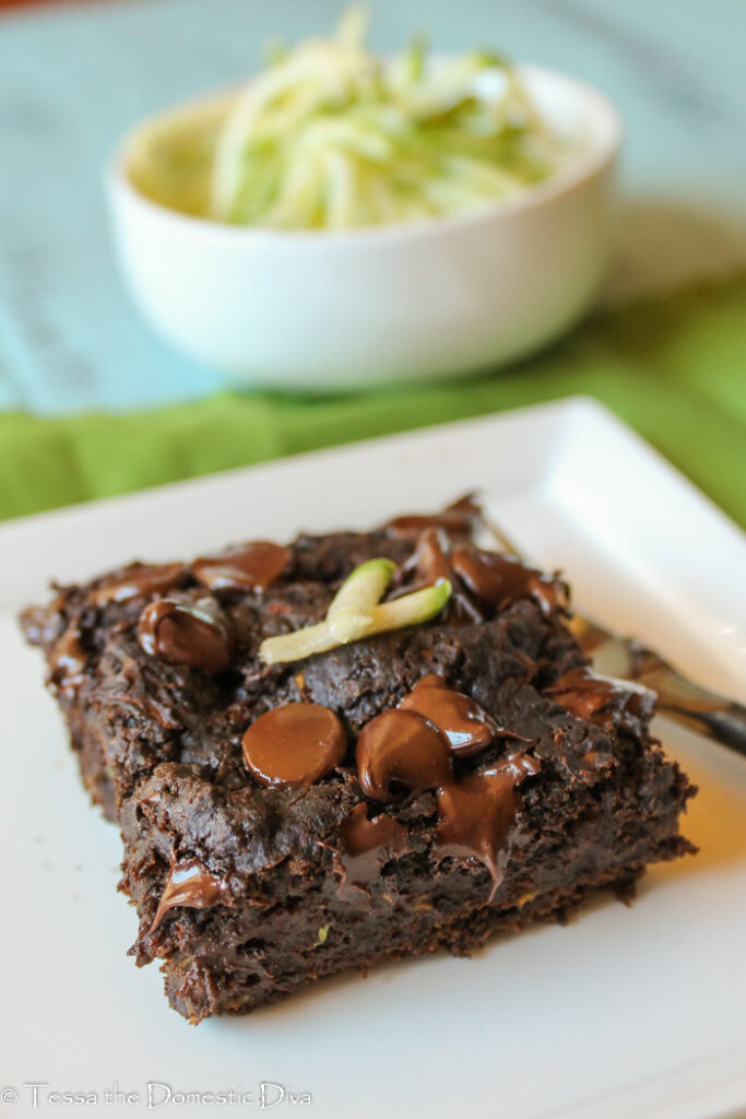 dark chocolate square of zucchini cake with melty chocolate chips and a garnish of fresh zucchini on a white plate