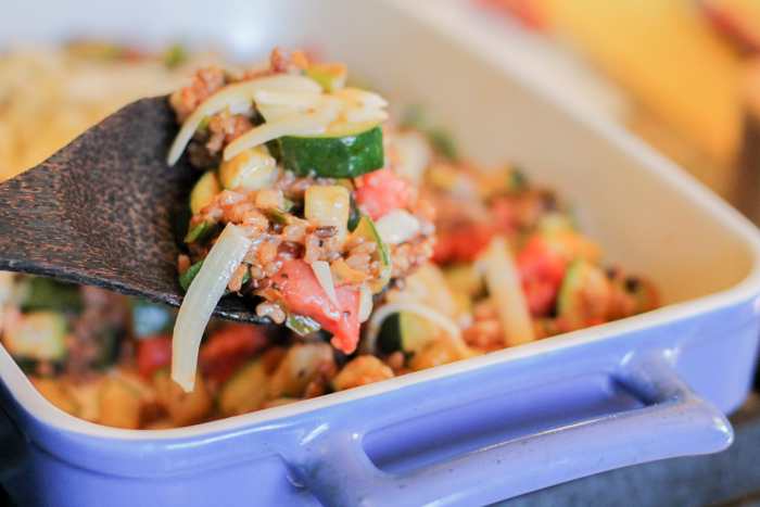 a blue ceramic baking pan with handles filed with brown rice, zucchini, tomotes, basil, and cheese and a serving spoon of coconut wood scooping up a serving