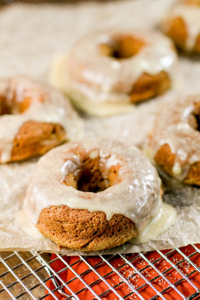 front view of glazed pumpkin donuts on top of a stainless steel cooling rack with a autumn hued orange fabric underneath