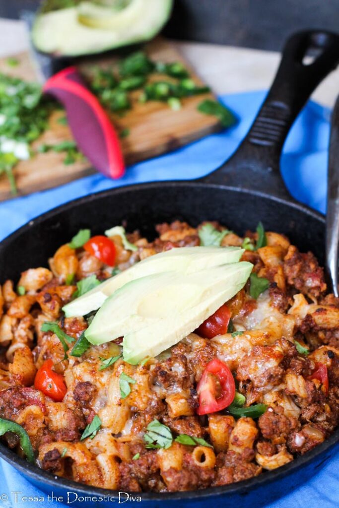 a pan filled with noodles, ground beef, and cheese topped with sliced avocado.