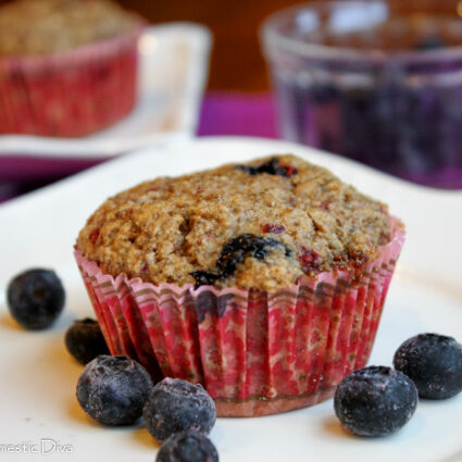 eye level close up of wholesome muffin studded with blueberries in a pink paper liner on white plate with fresh blueberries