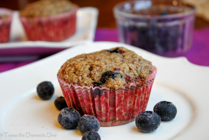 eye level close up of wholesome muffin studded with blueberries in a pink paper liner on white plate with fresh blueberries