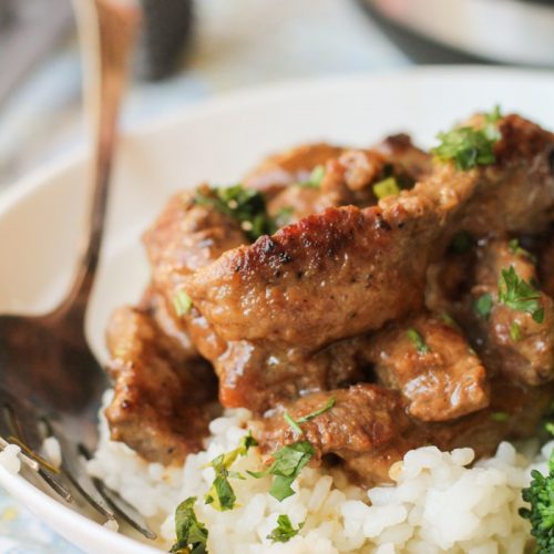 strips of tender steak coated in a savory gravy on top of a bed of white rice with a side of broccoli in a white bowl with an Instant Pot in the background