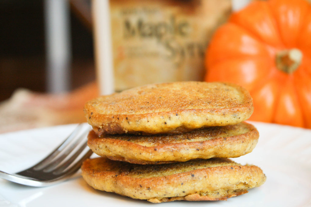 a horizontal image of three stacked golden orange pumpkin pancakes on a white plate with a fork