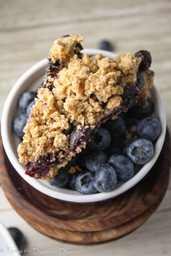 overhead shot of a triangle slice of crumbled oat topping and blueberry filled bar atop a bowl of fresh blueberries