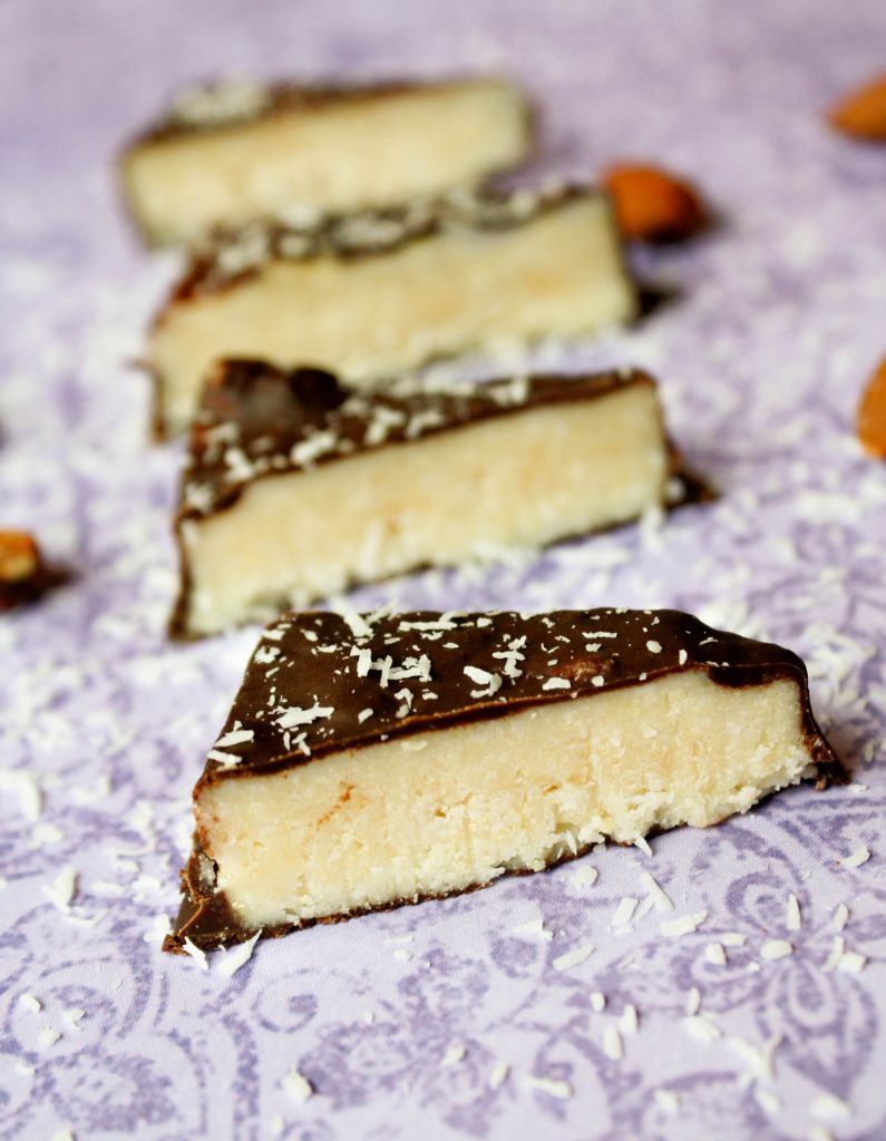 vertical image of keto almond joy bars coated in a sheen of low carb chocolate on a lavender fabric surrounded by whole almonds