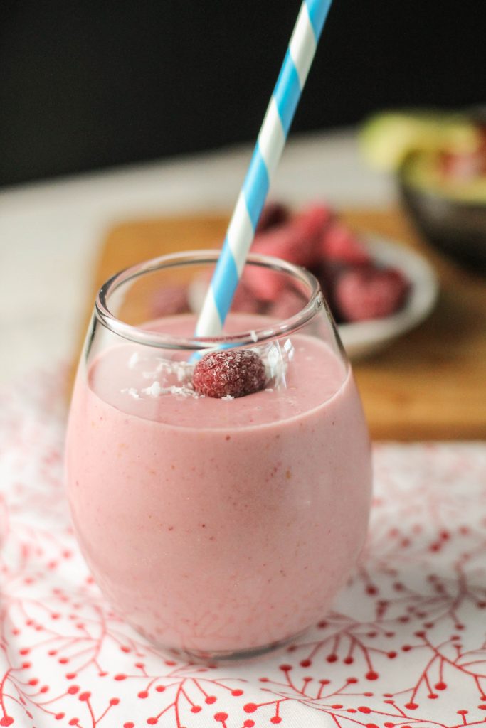 a clear glass filled with a creamy pink raspberry smoothie with avocado and fresh berries in background