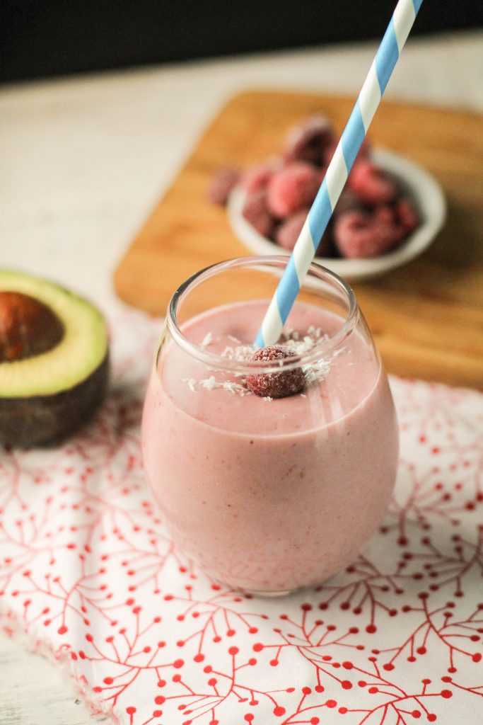 creamy plae pink raspberry smoothie garnihsed with white coconut flakes and a frozen raspberry in clear glass with a blute and white striped paper straw