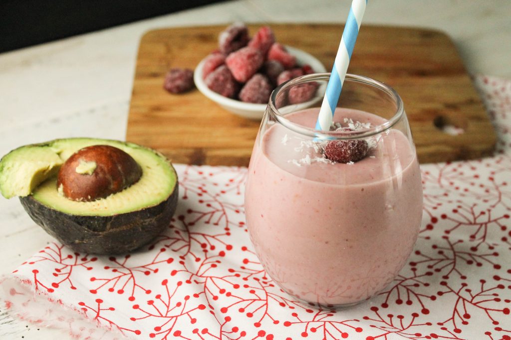 horizontal image of a pale pink raspberry smoothie in a clear glass with a blue and white paper straw, a halved avocado and a garnish of coconut flakes and frozen raspberries