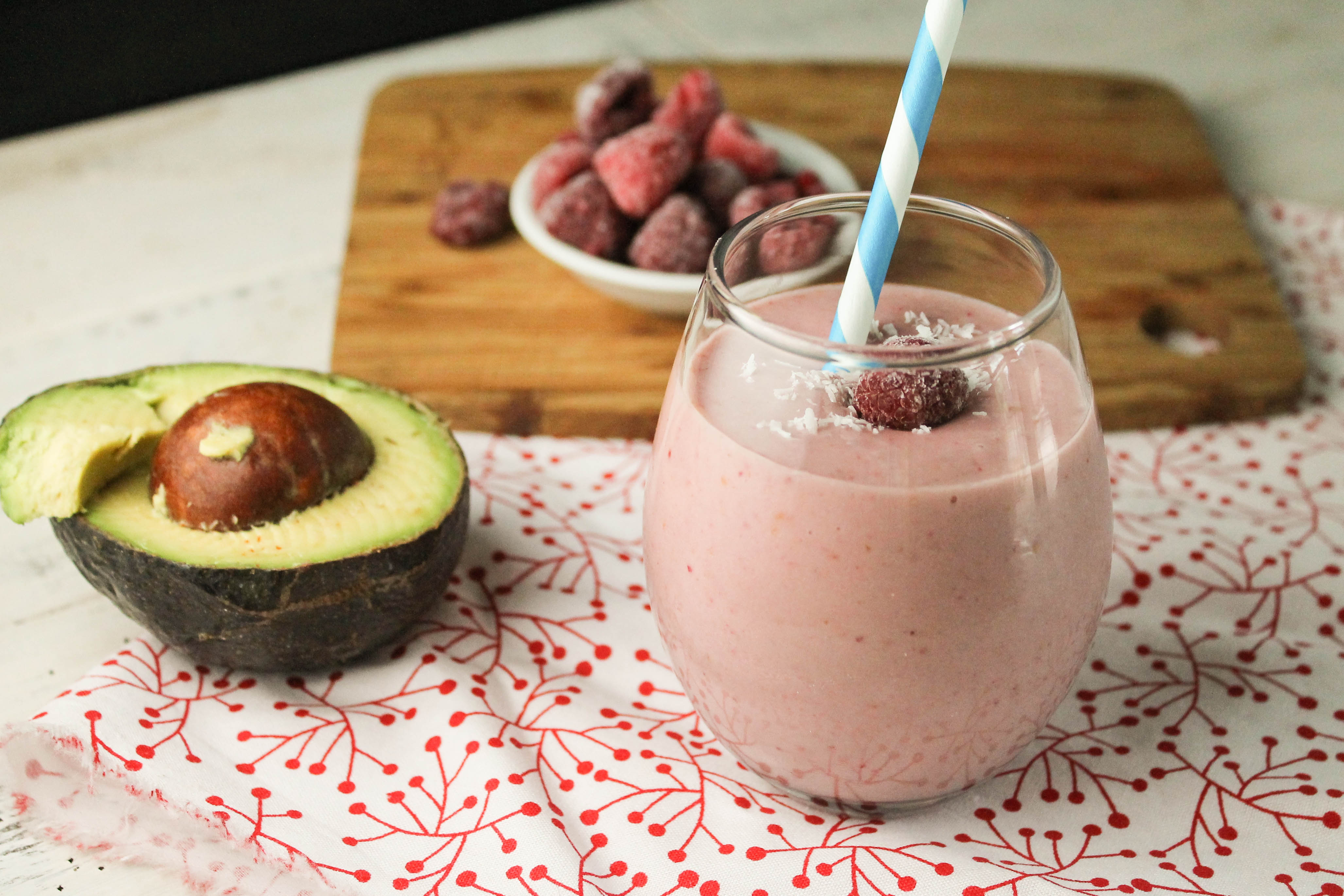 pale pink raspberry smoothie in a clear glass with a blue and white paper straw, a halved avocado and a garnish of coconut flakes and frozen raspberries