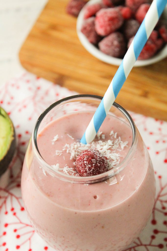 overhead shot of a pale pink creamy raspberry smoothie with a blue and white striped paper straw garnished with white coconut flakes and a frozen raspberry