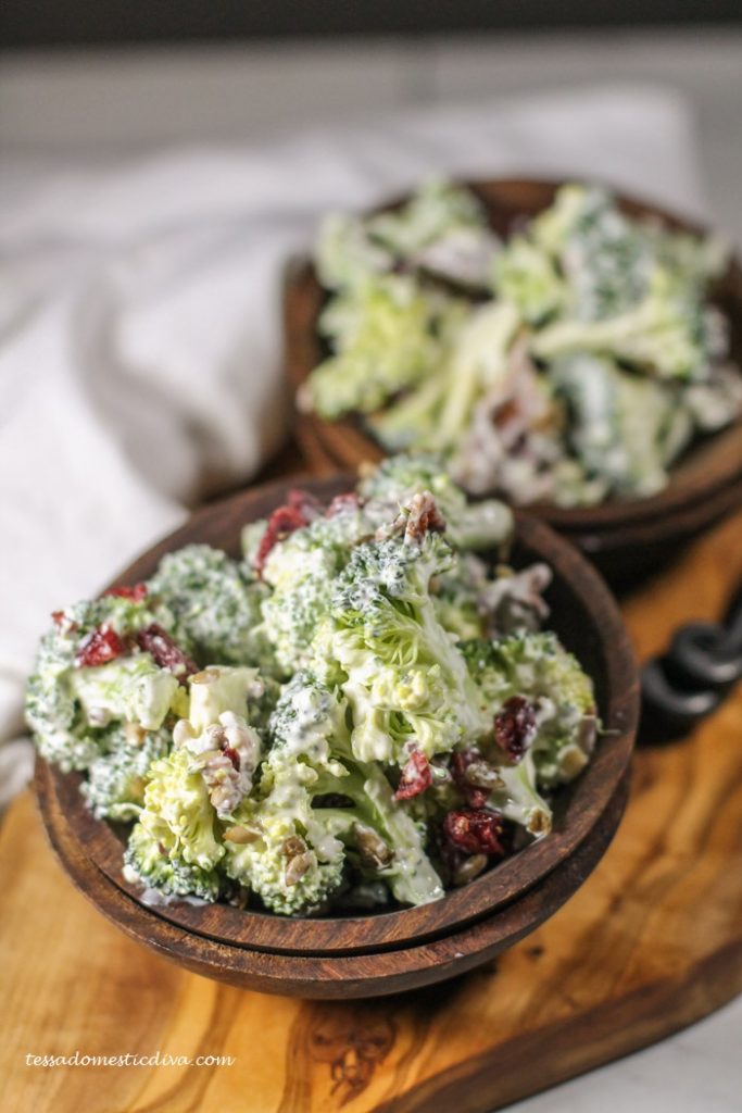 two deep brown wooden bowls filed with creamy broccoli bacon salad atop an olive wood cutting board