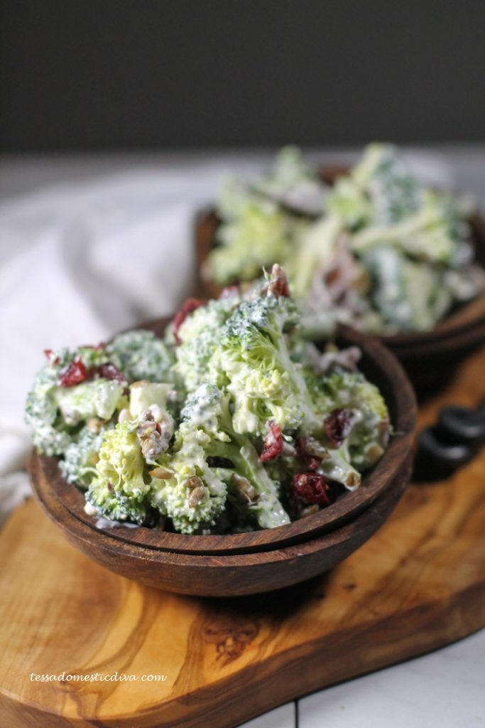 two wooden bowls filled with a creamy broccoli bacon salad with dots of bright red cranberries, aranged atop an live wood cutting board