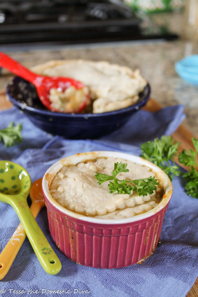 an individual ramekin with a buttery pastry topped root vegetable pot pie with fresh parsley and hand painted ceramic spoons