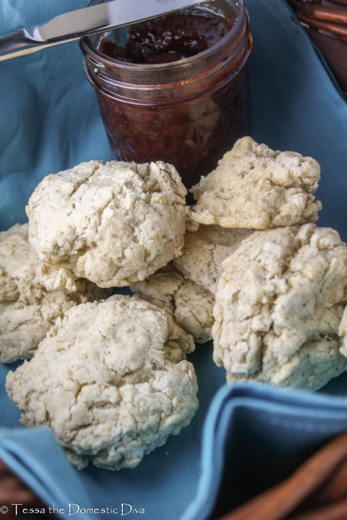 multiple biscuits arranged in a basket with a blue napkin and a jar of preserves