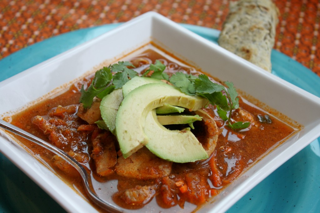 a square white bowl filled with a tomato and chicken soup with mexican spices and a topping of a avocado slices and cilantro