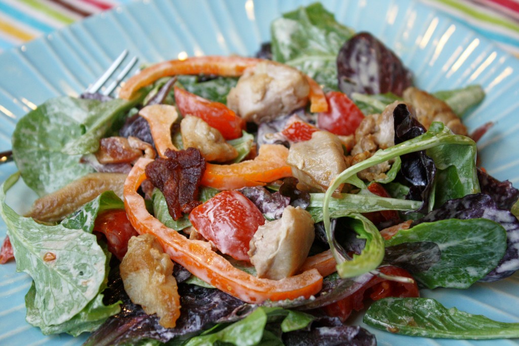 dressed mixed green salad topped with crispy bacon, browned chicken, red bell peppers, and tomatoes all on a turquoise blue plate with a festive striped cloth